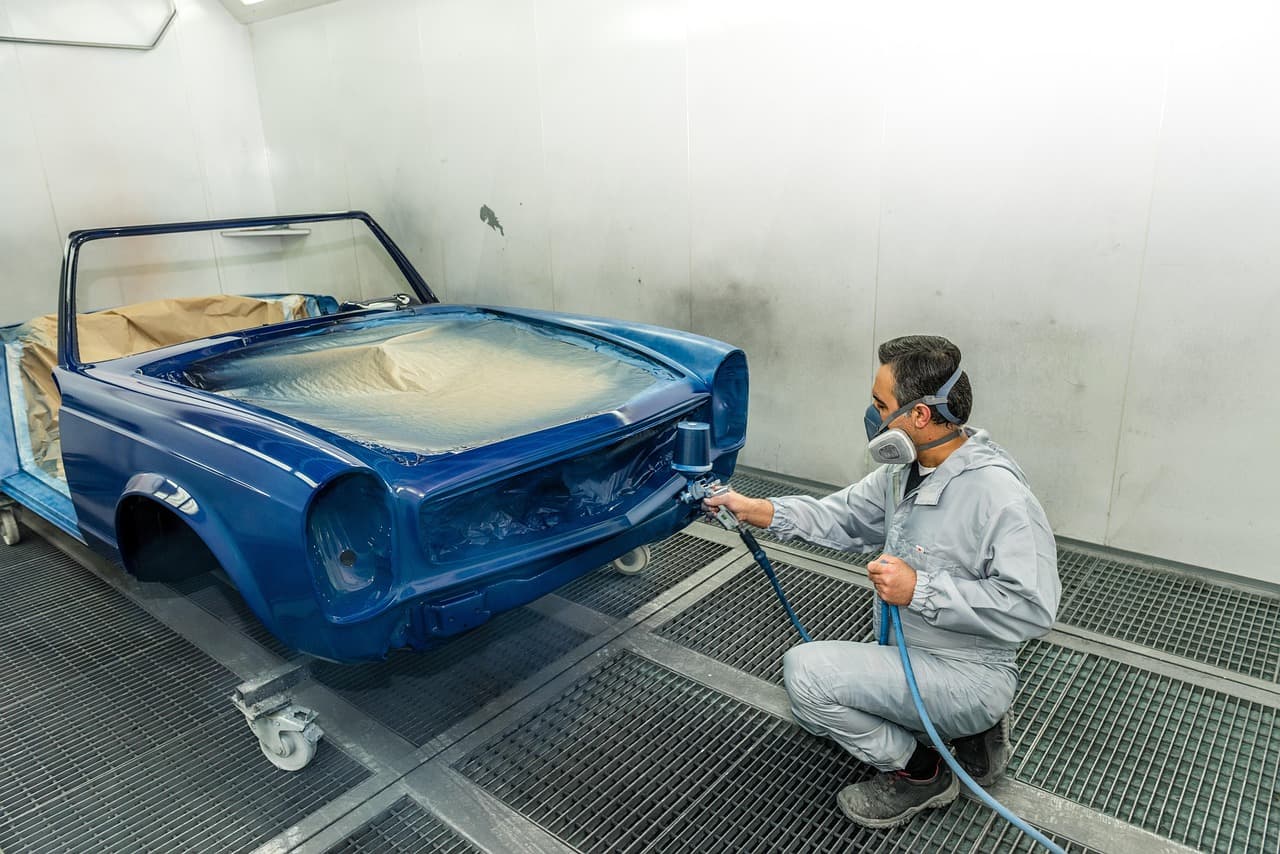 A painter in a mask spraying a blue car body in a white spray booth